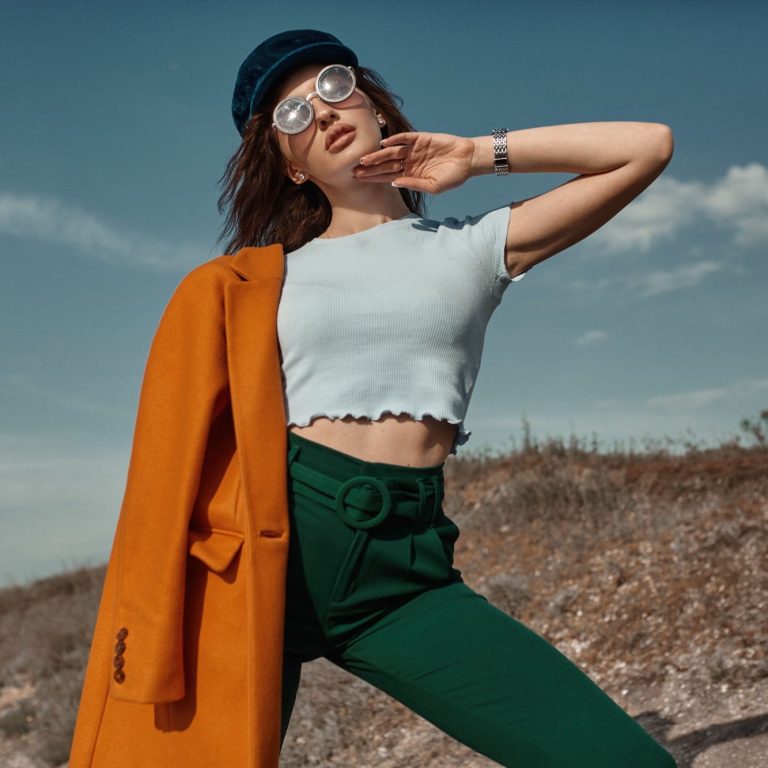 Femme élégante en manteau orange et pantalon vert, posant avec un chapeau et des lunettes.