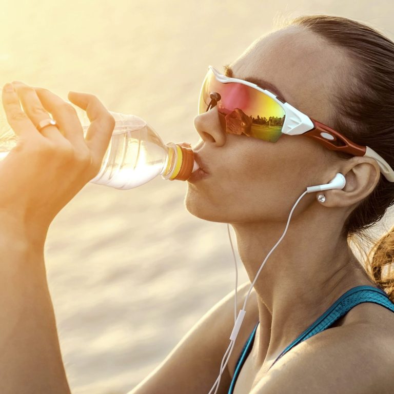 Femme buvant de l'eau, portant des lunettes de soleil et des écouteurs, avec un arrière-plan lumineux.