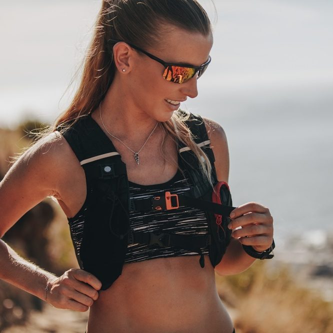 Femme souriante en tenue de sport, portant des lunettes de soleil sur un sentier en plein air.