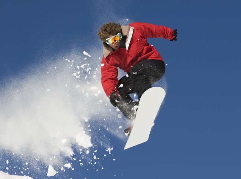 Un snowboarder en plein saut, projetant de la neige, sous un ciel bleu.